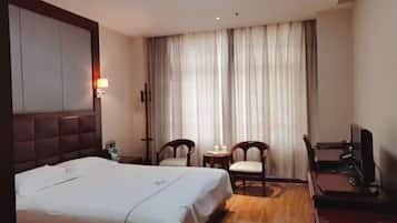 Habitación de hotel con cama, dos sillas, una mesita, televisor y un ventanal con cortinas.