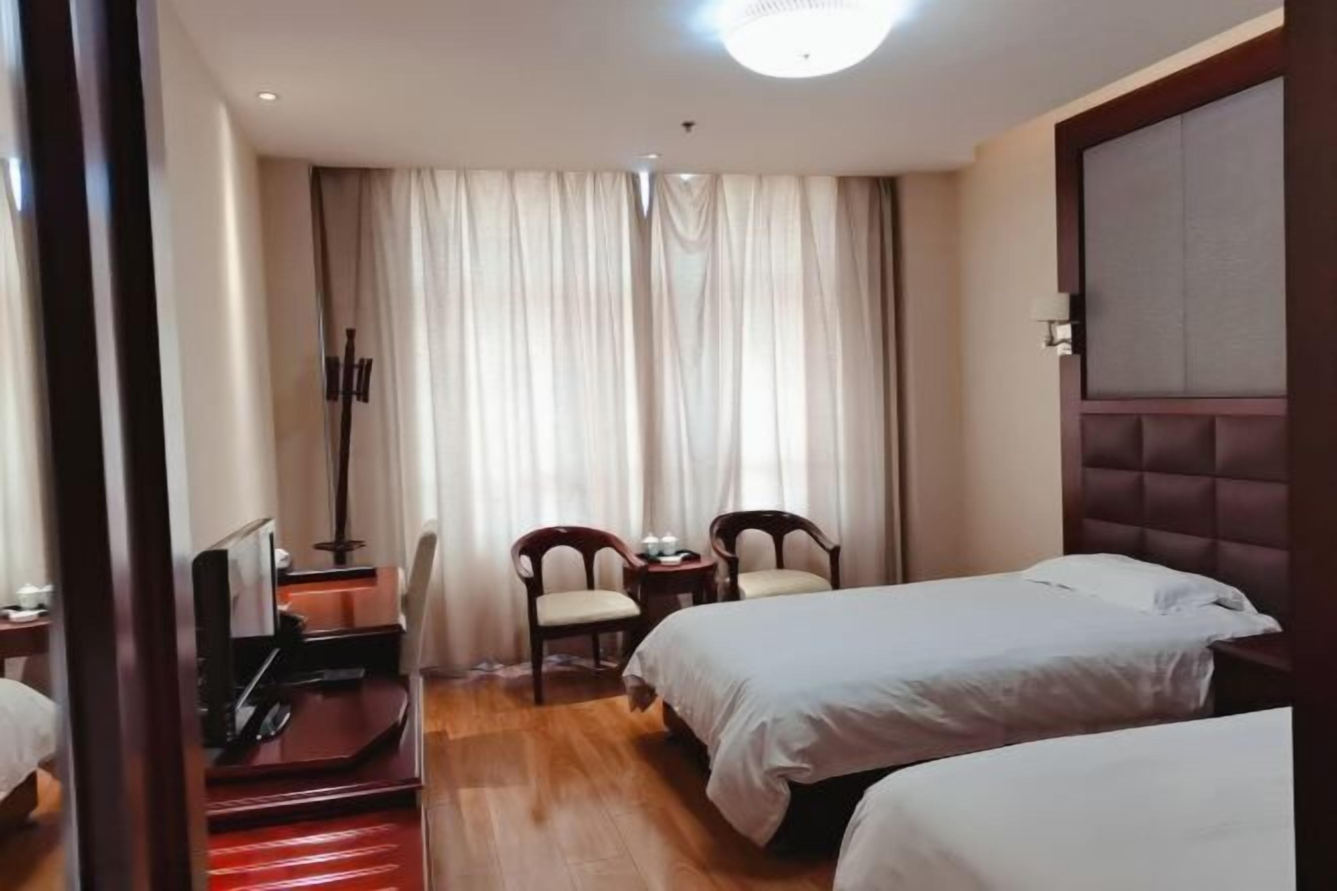 A hotel room with two beds, a desk with a chair, a small table with two chairs, and a large window with curtains.