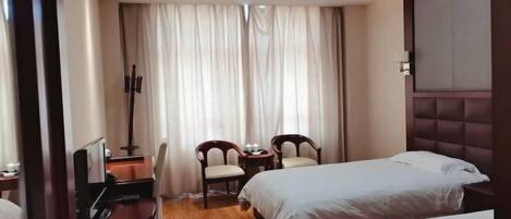 A hotel room with two beds, a desk with a chair, a small table with two chairs, and a large window with curtains.