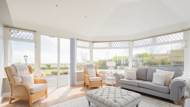 A spacious living room with a grey sofa, two wicker chairs, a grey ottoman, and a white coffee table. Large glass doors lead to a garden. - Sea Haven - Stunning Sea Views! (County Clare)