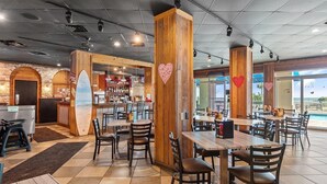 En restaurang med träpelare, en surfbräda som dekoration och utsikt över stranden.