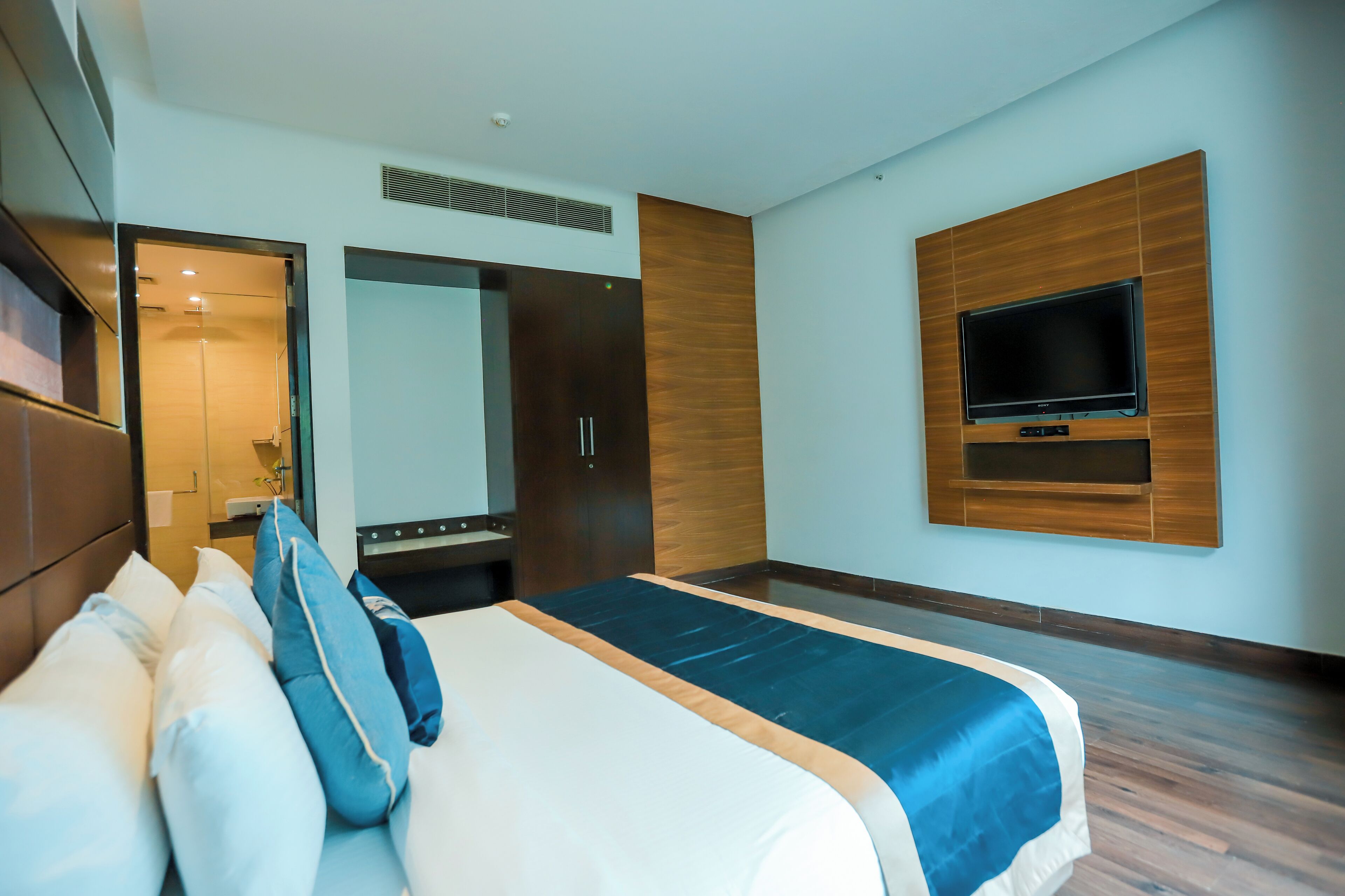 A modern hotel room with a large bed, a TV mounted on the wall, and a mirror above the bed.