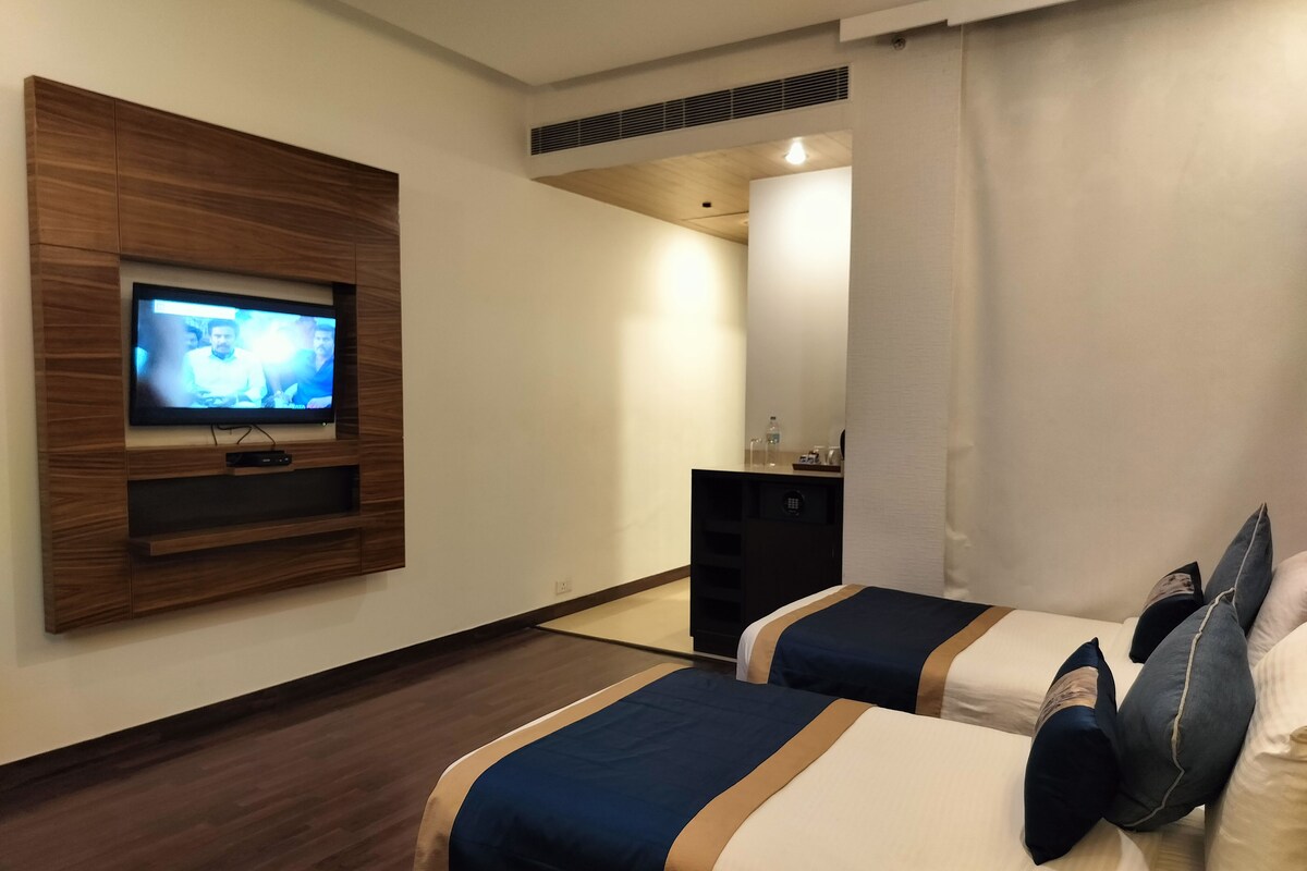 A hotel room with two beds, a TV mounted on a wooden panel, a desk with a chair, and a window with curtains.