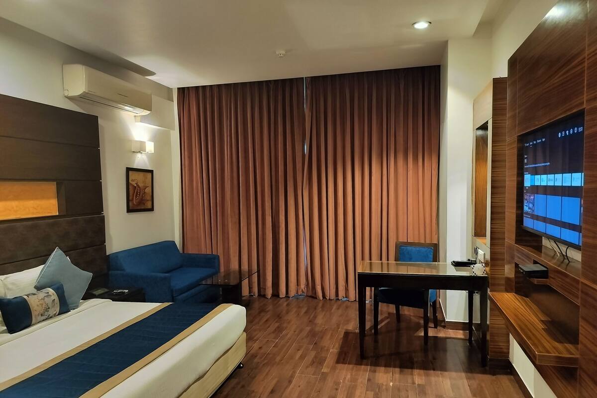 Una habitación de hotel moderna con una cama grande, un sofá azul, una mesita pequeña, un televisor montado en la pared y una ventana con cortinas.