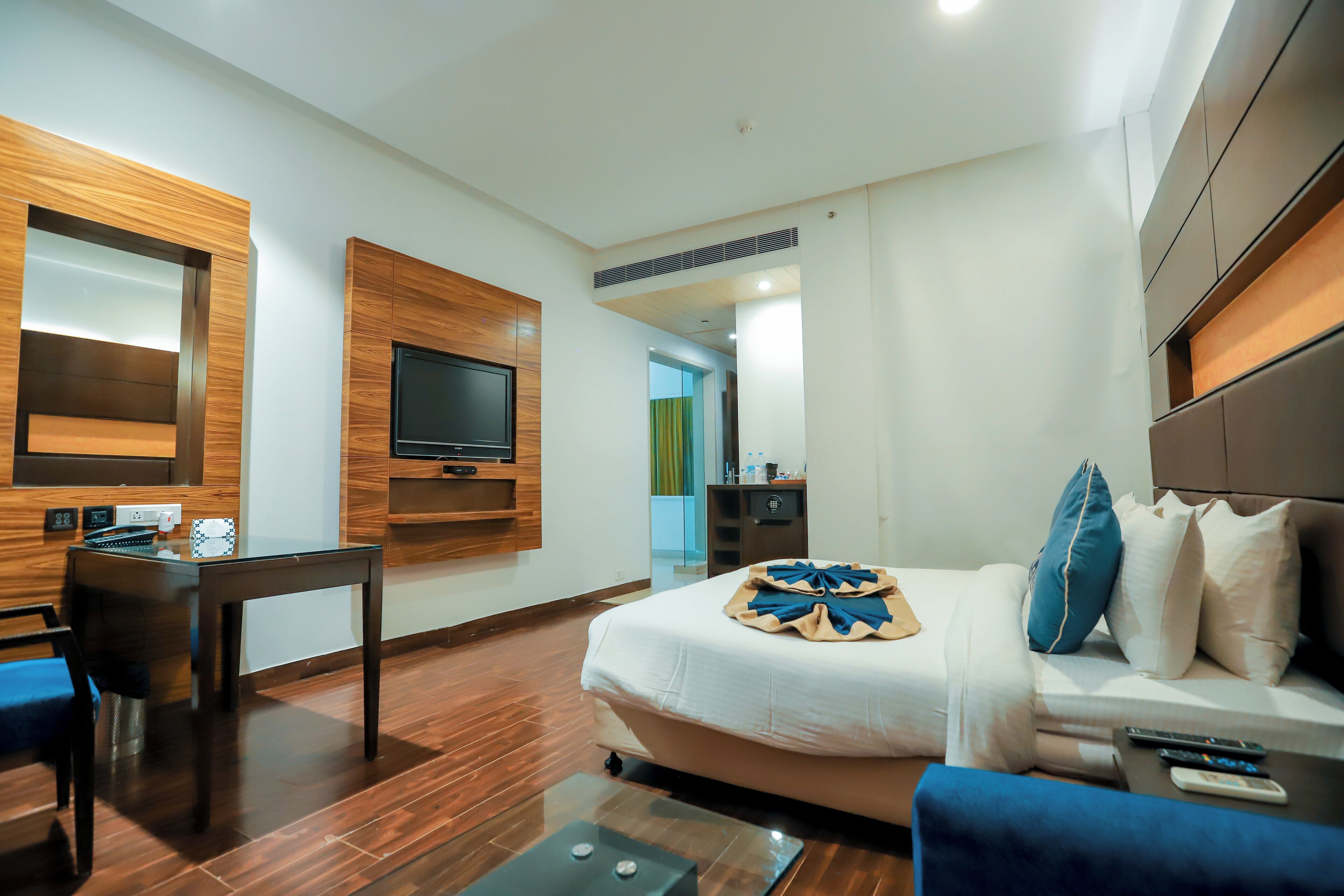 A modern hotel room with a large bed, a TV mounted on the wall, a small table, and a seating area with a blue sofa.