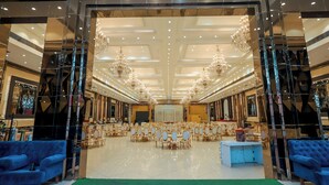A spacious banquet hall with rows of round tables and chairs, a stage at the front, and chandeliers. - Bel-La Monde Hotel Chhattarpur South Delhi (New Delhi)