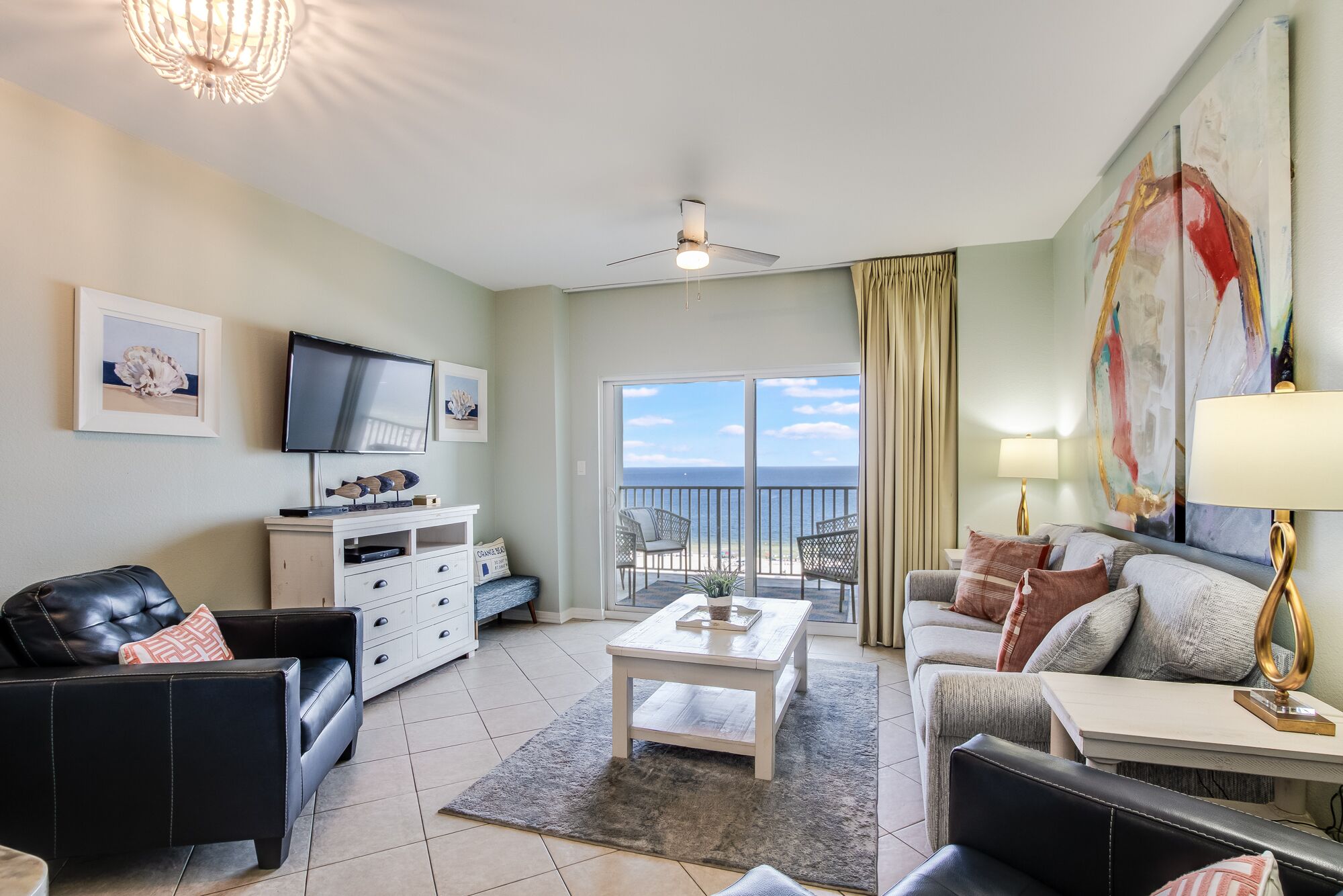 A spacious living room with a television, a white cabinet, a coffee table, and a sofa. There is a balcony with a view of the ocean.