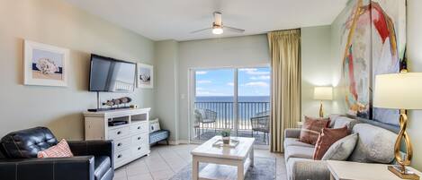 A spacious living room with a television, a white cabinet, a coffee table, and a sofa. There is a balcony with a view of the ocean.