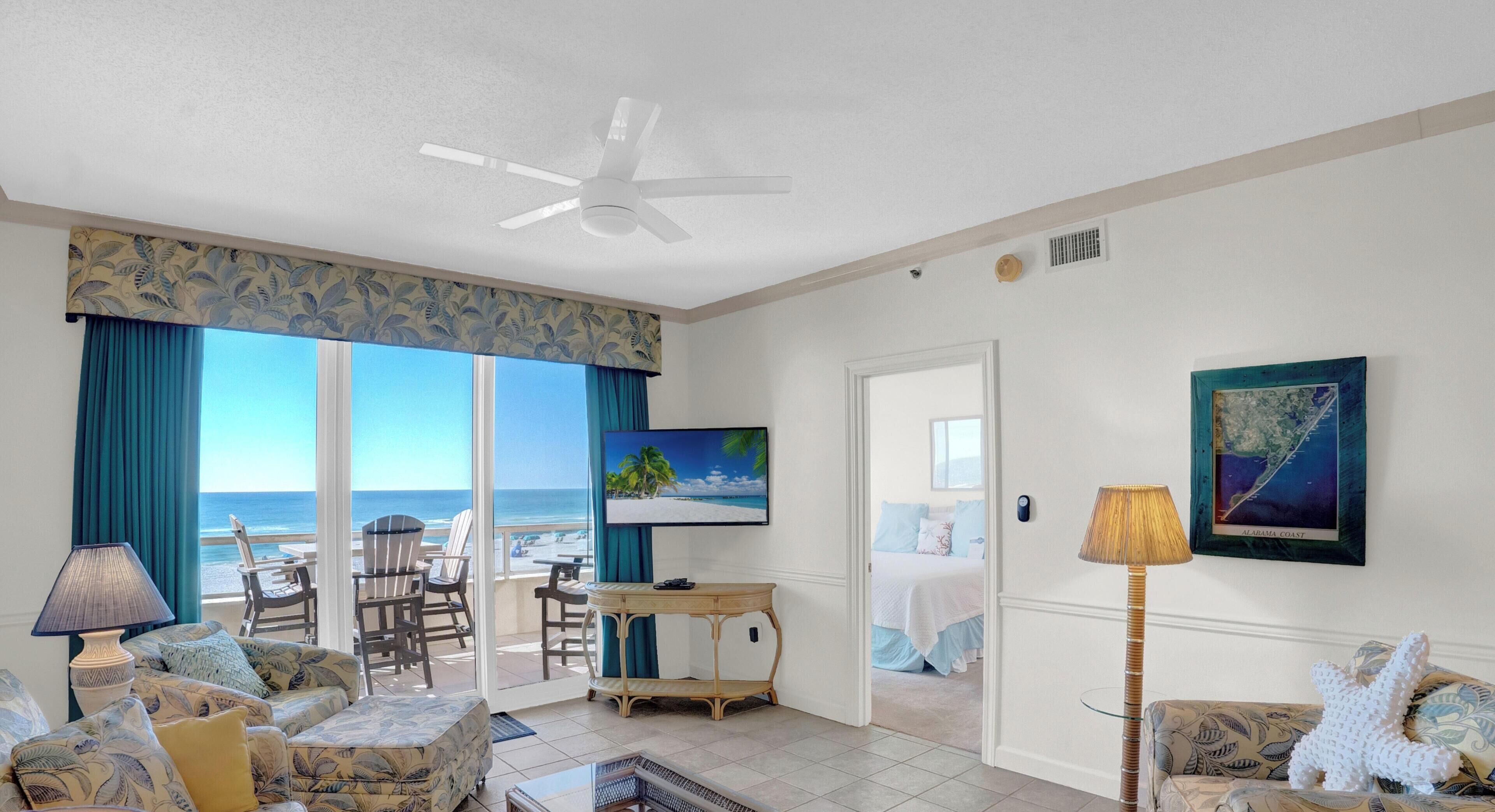 A room with a view of the ocean, a ceiling fan, a television, a bed, and a table.
