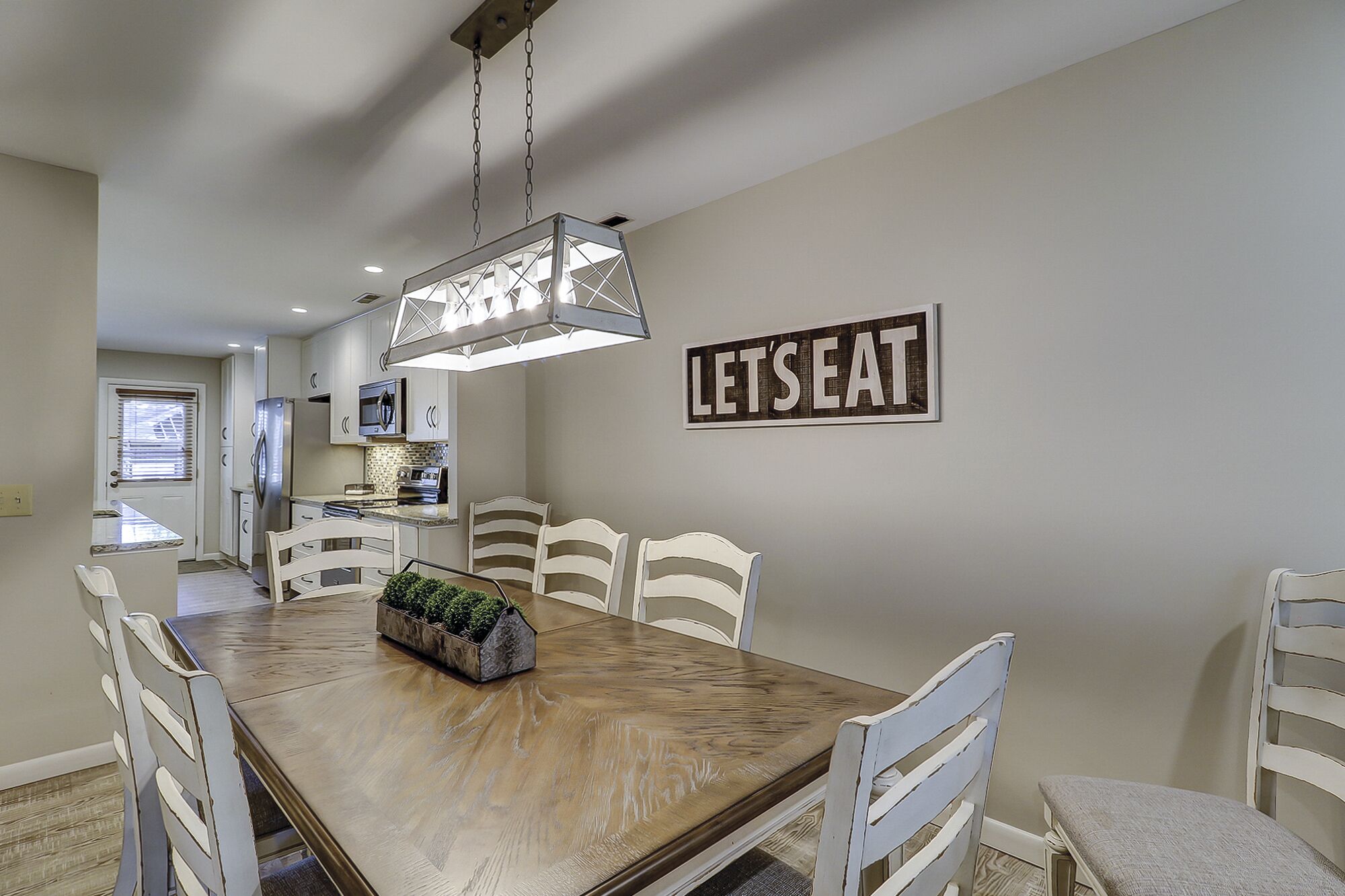A dining area with a wooden table, white chairs, a hanging light fixture, and a 'LET'S EAT' sign on the wall.