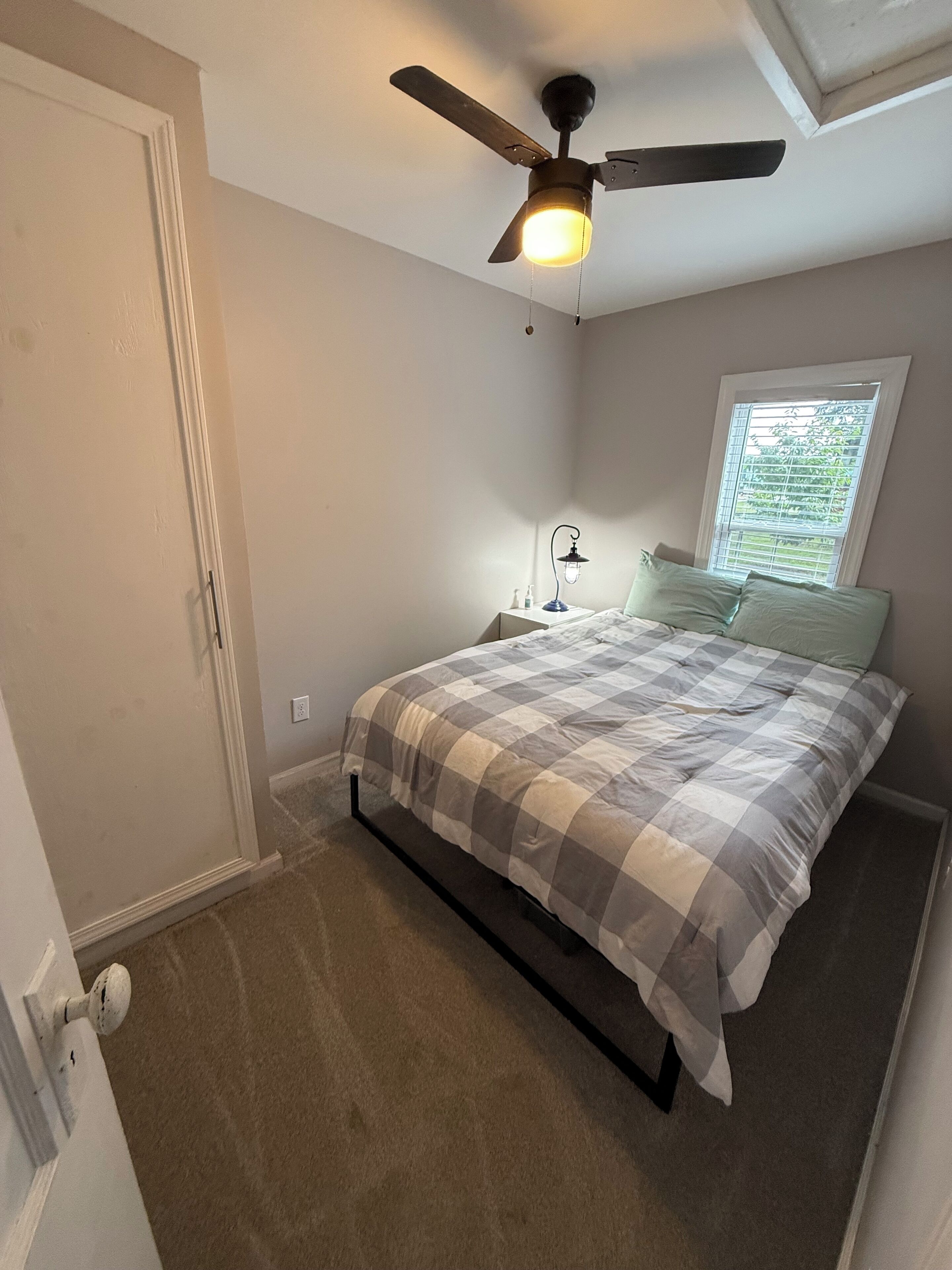 A bedroom with a bed, a ceiling fan, a nightstand, a window, and a door.