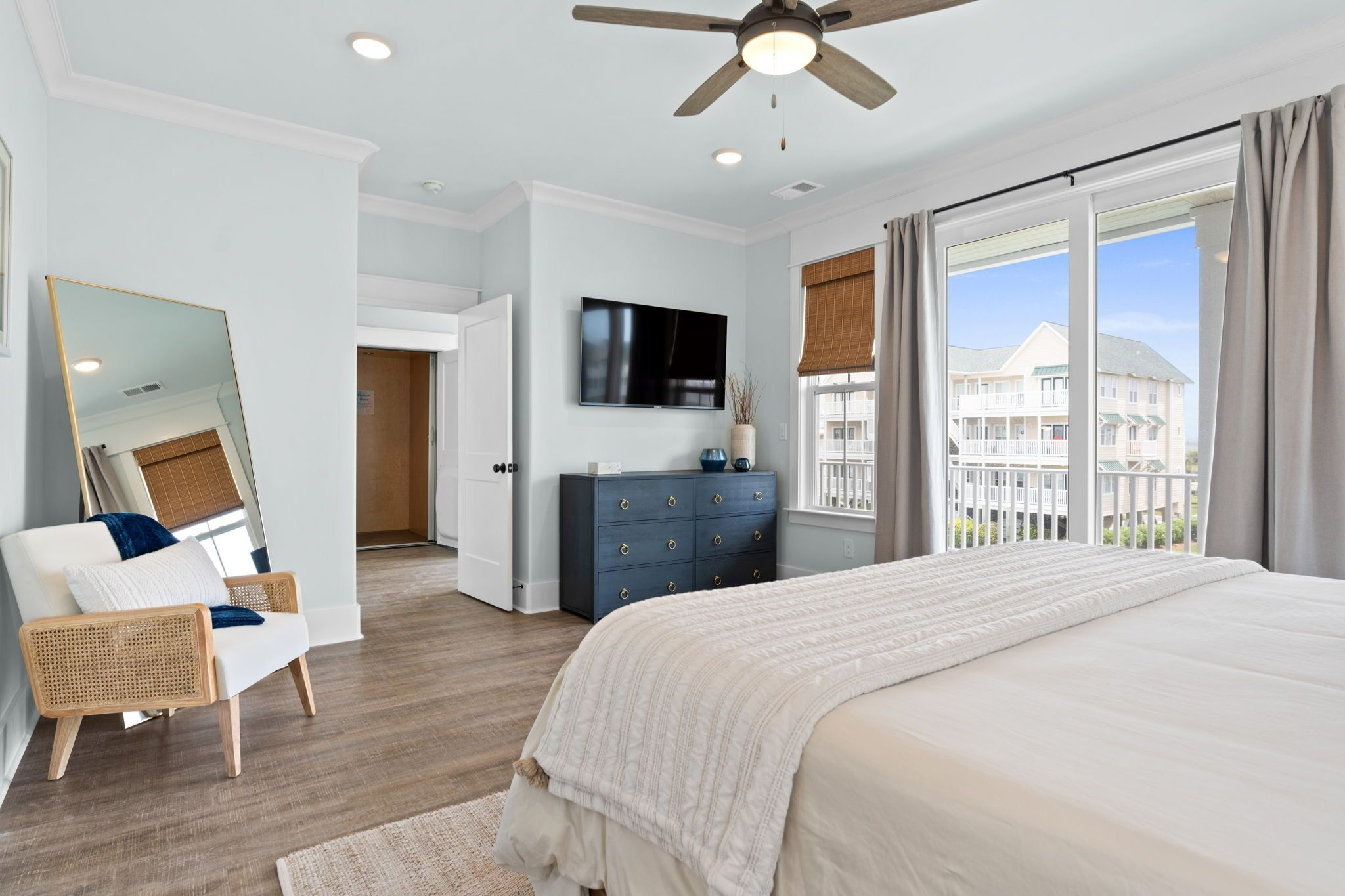 A bedroom with a large bed, a ceiling fan, a TV, a dresser, and a window with a view of buildings.