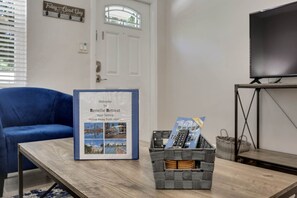 A framed welcome sign for a retreat, a blue chair, a wooden table, a basket with magazines, and a TV on a stand.