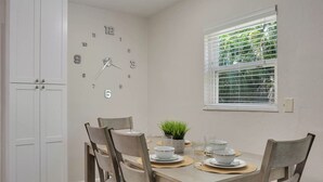 Un coin repas avec une horloge au mur, une fenêtre avec des stores et une table mise avec des assiettes et des bols.