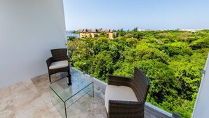 A balcony with wicker furniture and a glass table, overlooking a lush green landscape and a building with a thatched roof. - Dream 2BR PH! Steps to Beach! Downtown location! (Playa del Carmen)