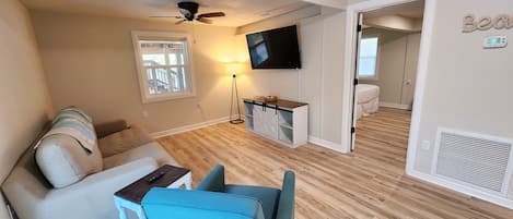 A living room with a sofa, a blue chair, a TV mounted on the wall, a ceiling fan, and a small table with a remote control.