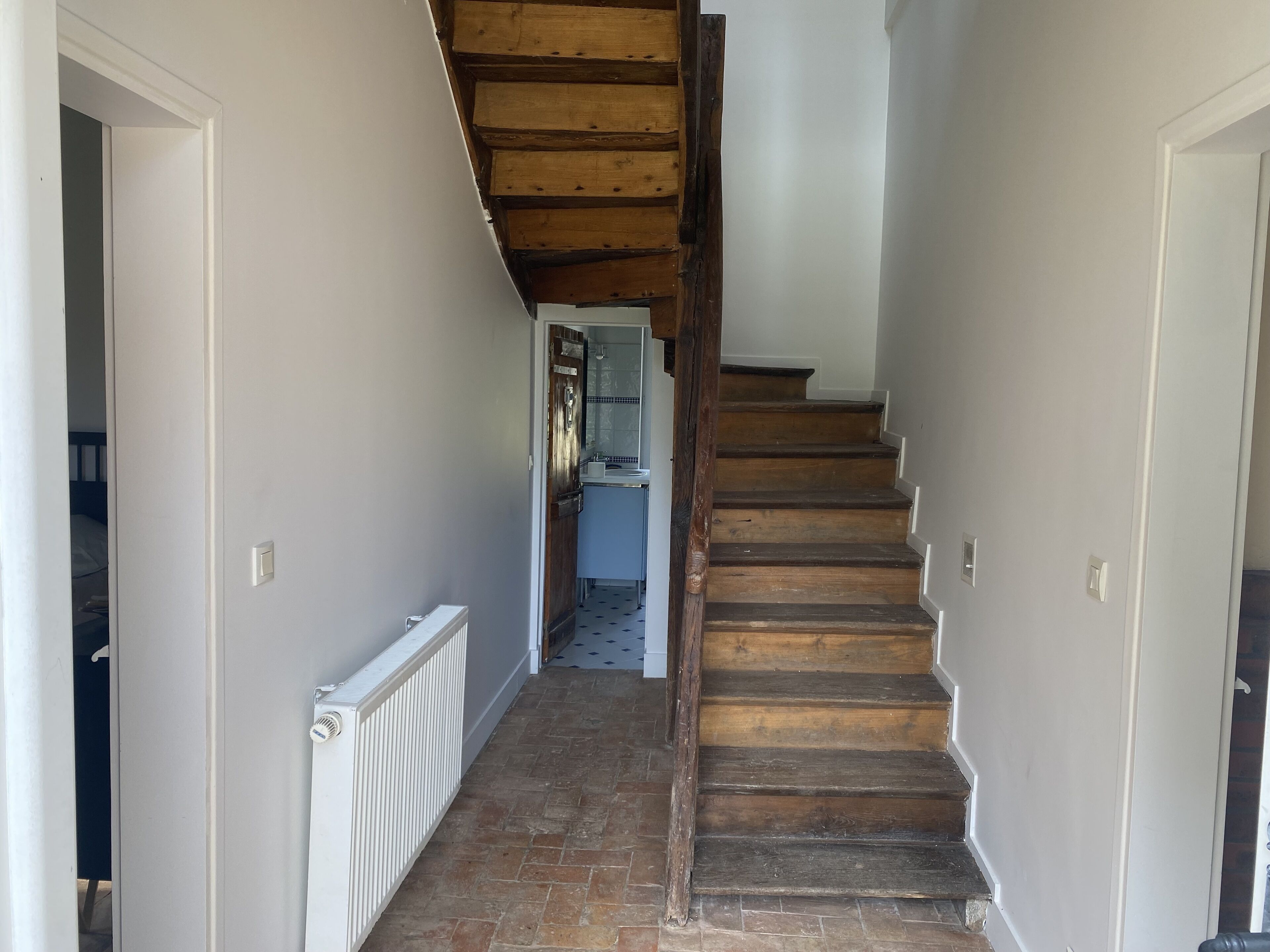 A narrow hallway with a wooden staircase leading to a kitchen area.