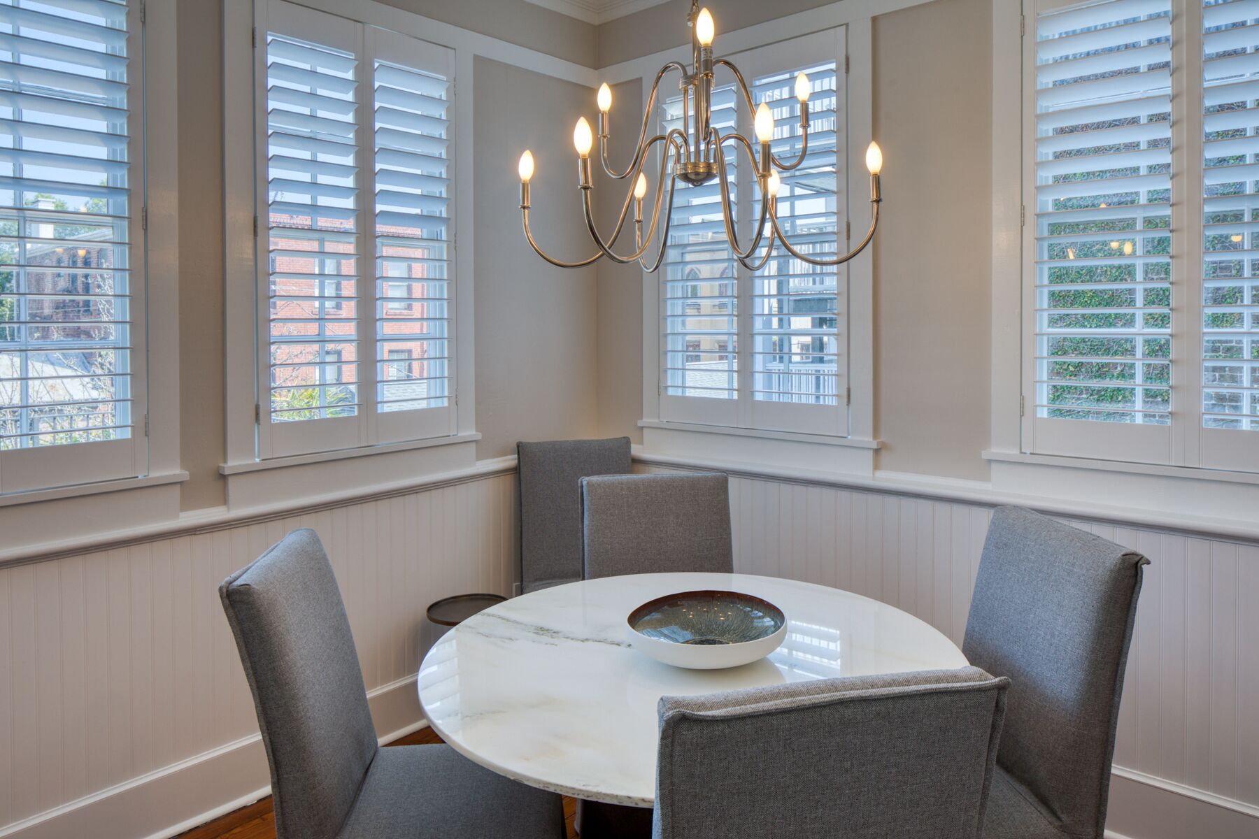 A dining area with a round table, grey upholstered chairs, and a chandelier.
