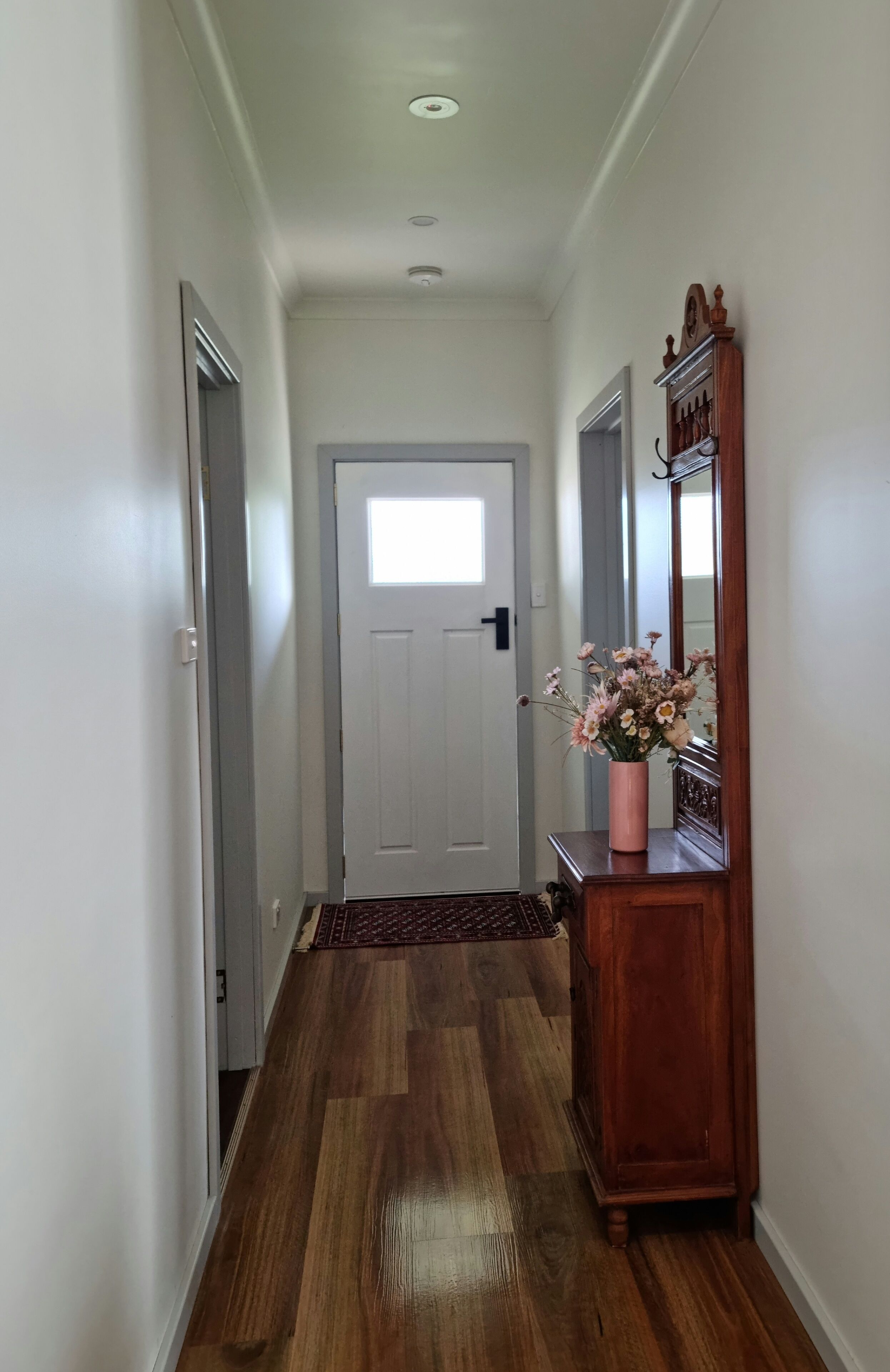 Un pasillo con suelo de madera, una puerta blanca al final y una consola de madera con un jarrón de flores.