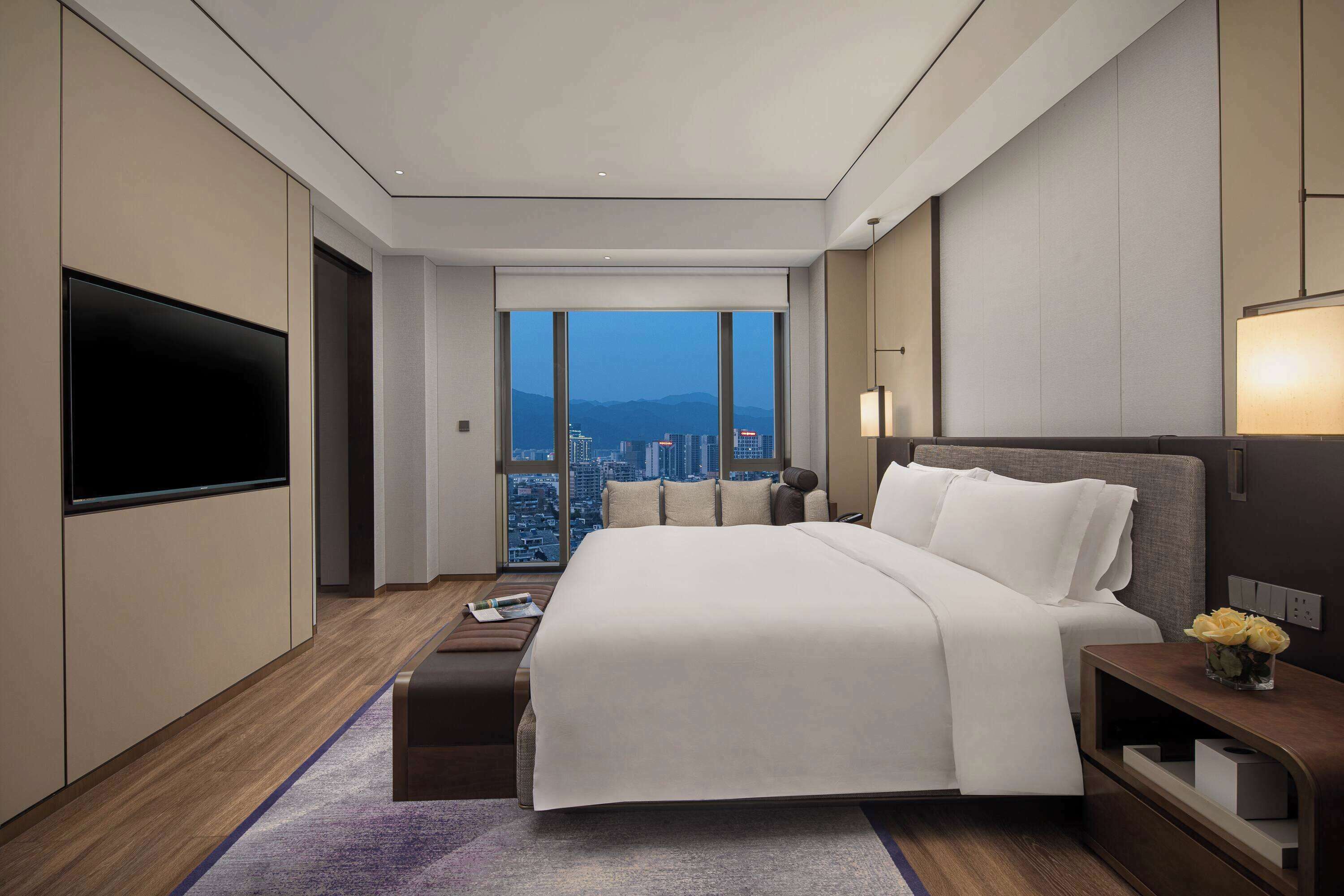 Habitación de hotel moderna con una cama grande, un televisor de pantalla plana, vistas a la ciudad y una mesita de noche con un jarrón de flores.