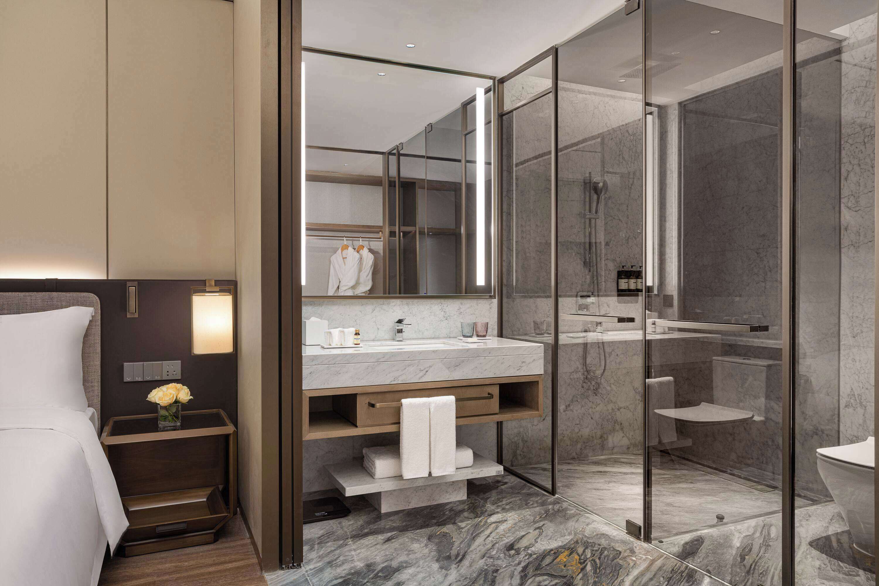 Une salle de bain moderne avec une douche à parois de verre, un lavabo en marbre et une table de chevet agrémentée d’un vase de fleurs.