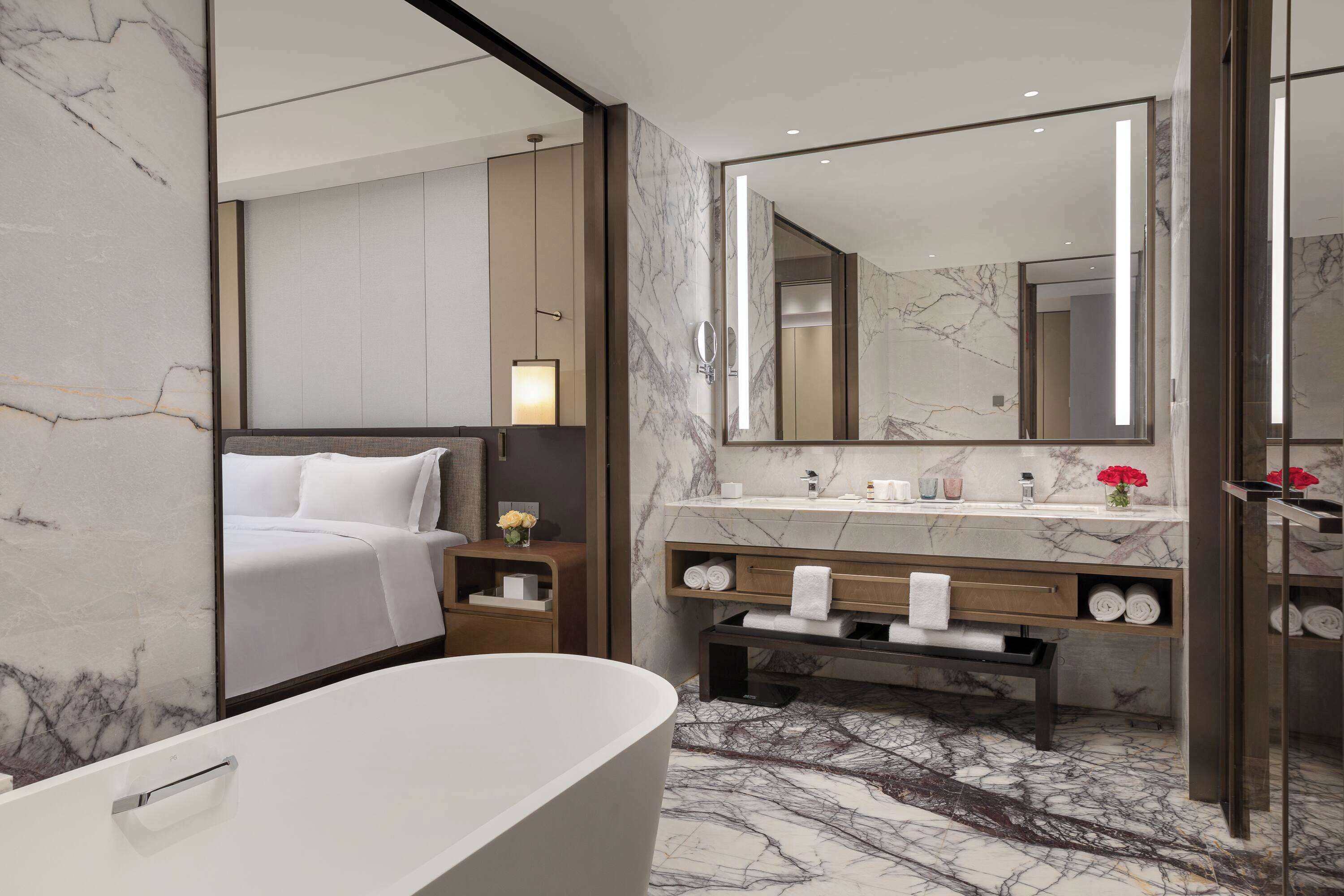 A modern bathroom with a marble floor, a large mirror, a bathtub, a vanity with a marble countertop, and a bedside table with a lamp.