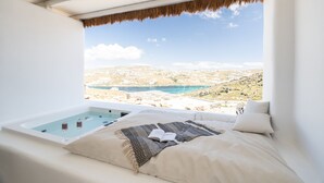 A bedroom with a large window overlooking a scenic landscape, a bathtub, and a bed with a blanket. - White Stone Mykonos Hotel (Mykonos)