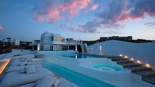 A modern pool area with lounge chairs and a hot tub, surrounded by a white building with a view of the sea. - White Stone Mykonos Hotel (Mykonos)