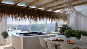 A modern kitchen with a large island, a dining area with wicker chairs, and a view of a cityscape through large windows. - White Stone Mykonos Hotel (Mykonos)