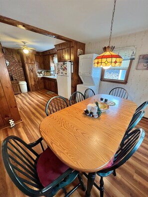 A wooden dining table with chairs, a hanging light fixture, and a kitchen area with wooden cabinets and appliances. - Waterfront on Sebago Lake (Standish)