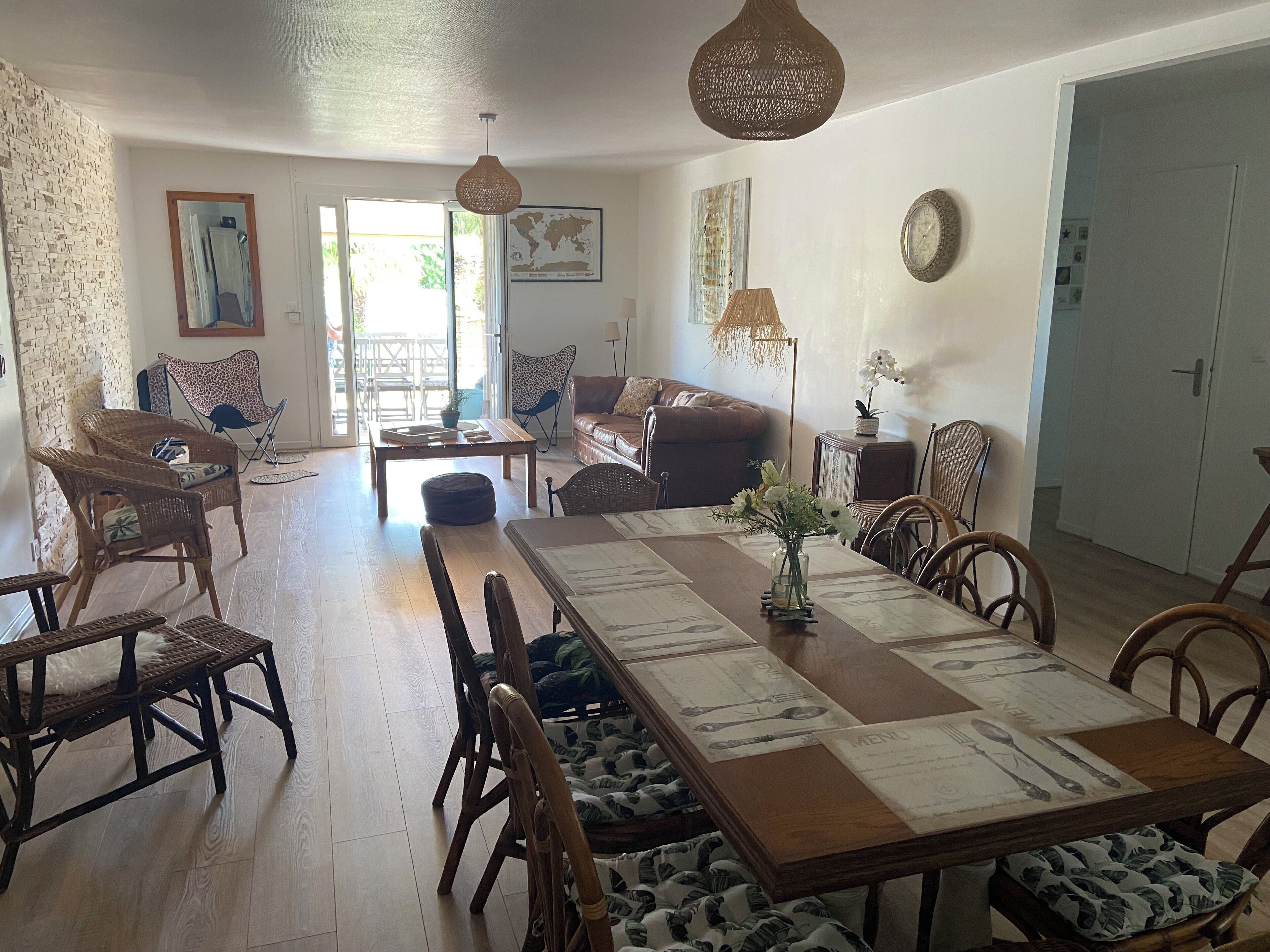 A dining area with a long wooden table, chairs, and a vase of flowers. There is a sofa and a chair in the background, and a door leading to another room.