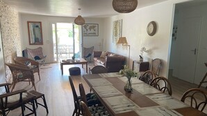 Un coin repas avec une longue table en bois, des chaises et un vase de fleurs. Au fond, on aperçoit un canapé et un fauteuil, ainsi qu’une porte donnant sur une autre pièce.