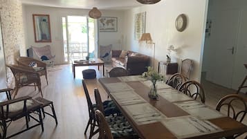 A dining area with a long wooden table, chairs, and a vase of flowers. There is a sofa and a chair in the background, and a door leading to another room.