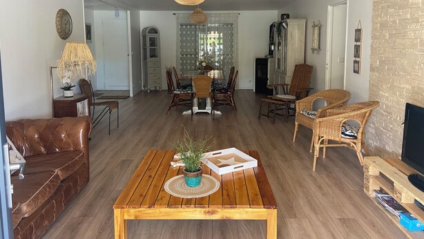 A spacious living room with a wooden coffee table, wicker chairs, a brown leather sofa, and a dining area with a wooden table and chairs.
