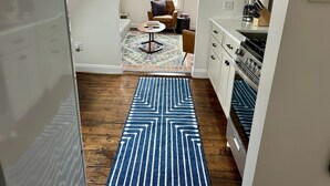 A modern kitchen with white cabinets and a built-in oven. A blue patterned rug leads to a living area with a patterned rug and a brown armchair.