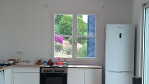 Une cuisine avec des meubles blancs, un four noir, un réfrigérateur blanc et une fenêtre avec des volets bleus.