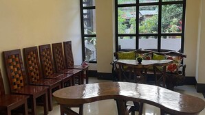 Uma área de refeições com uma mesa de madeira comprida e cadeiras, janelas amplas com vista para vegetação e teto com iluminação embutida.