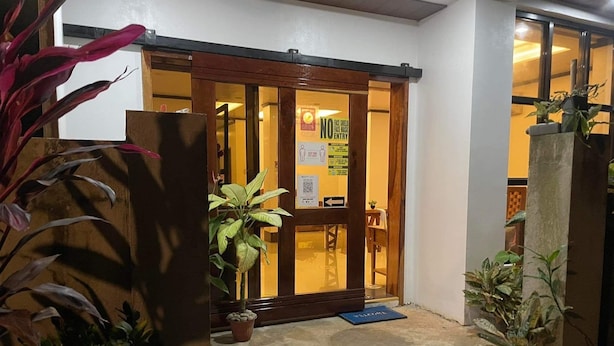 Entrada de hotel com porta de madeira, placa com a inscrição 'Obrigatório uso de máscara facial, sem protetor facial' e vasos de plantas.