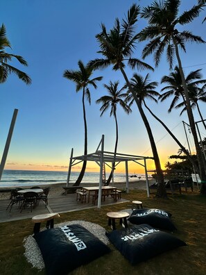 Un espace aménagé avec des tables et des bancs, situé au bord de la plage, entouré de palmiers et offrant une vue sur l’océan.