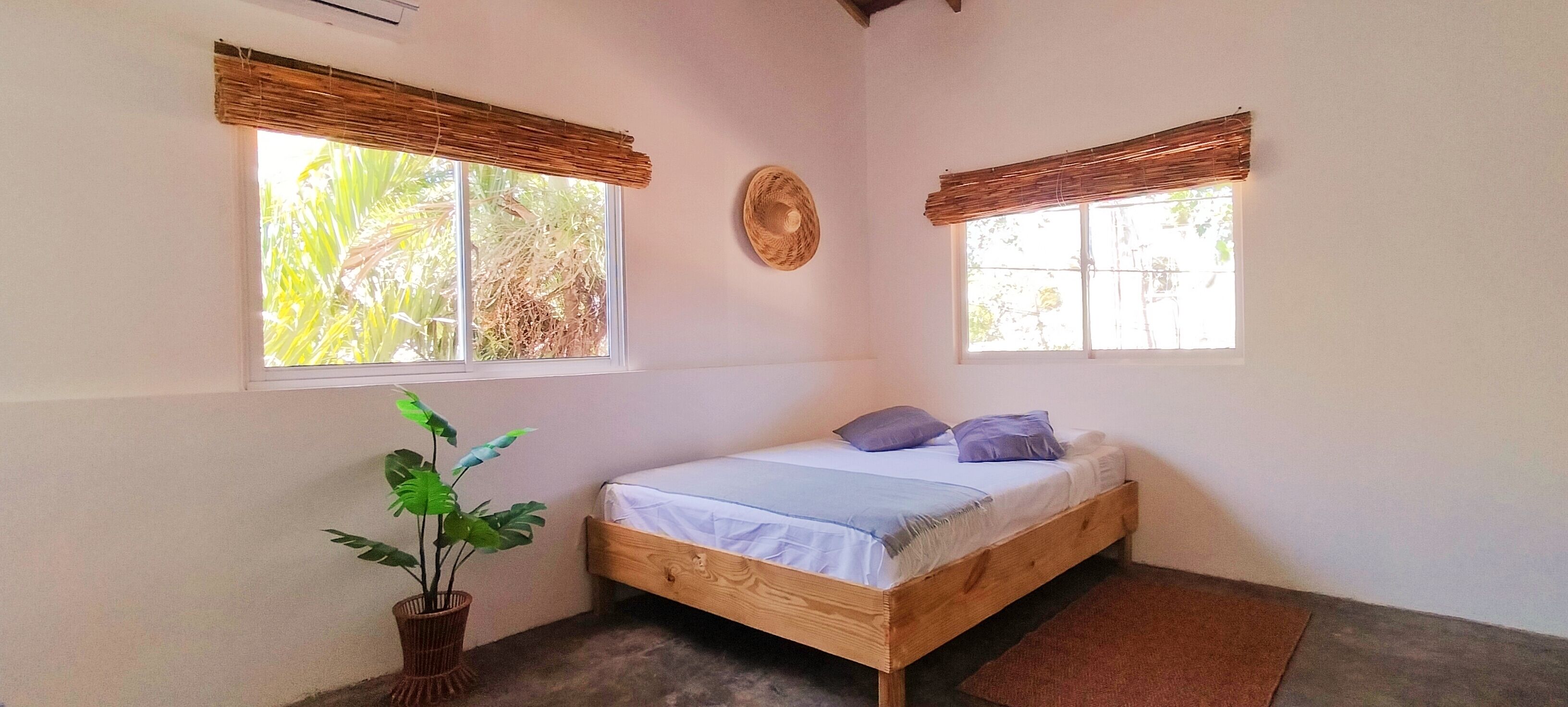 A small, single-bed room with a wooden frame, two windows with bamboo blinds, a potted plant, and a woven wall hanging.