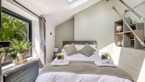 Un dormitorio con tragaluz, cama con ropa de cama blanca, mesita de noche con lámpara y vistas al exterior con vegetación.