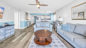 A living room with a round wooden coffee table, a grey sofa, a ceiling fan, and a kitchen in the background.