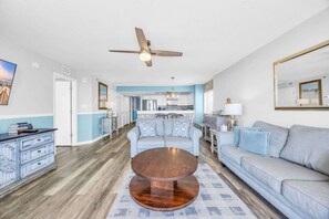 A living room with a round wooden coffee table, a grey sofa, a ceiling fan, and a kitchen in the background.