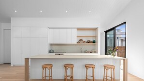 A modern kitchen with a central island, three bar stools, white cabinetry, and a large window. - Gorgeous George Barwon Heads (Barwon Heads)