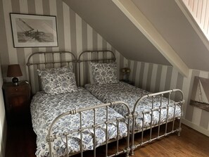 A bedroom with a sloped ceiling, a bed with a metal frame, a nightstand with a lamp, and a framed picture on the wall. - Charmante Maison Familiale (Binic-Étables-sur-Mer)