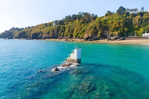 A coastal scene with a lighthouse on a rocky outcrop, clear turquoise waters, and a forested hillside with buildings. - Charmante Maison Familiale (Binic-Étables-sur-Mer)