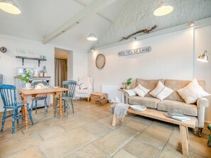 Un salon chaleureux avec une table en bois, un fauteuil à rayures et un canapé beige agrémenté de coussins. Une pancarte sur le mur indique : « NEW HOUSE LANE ».