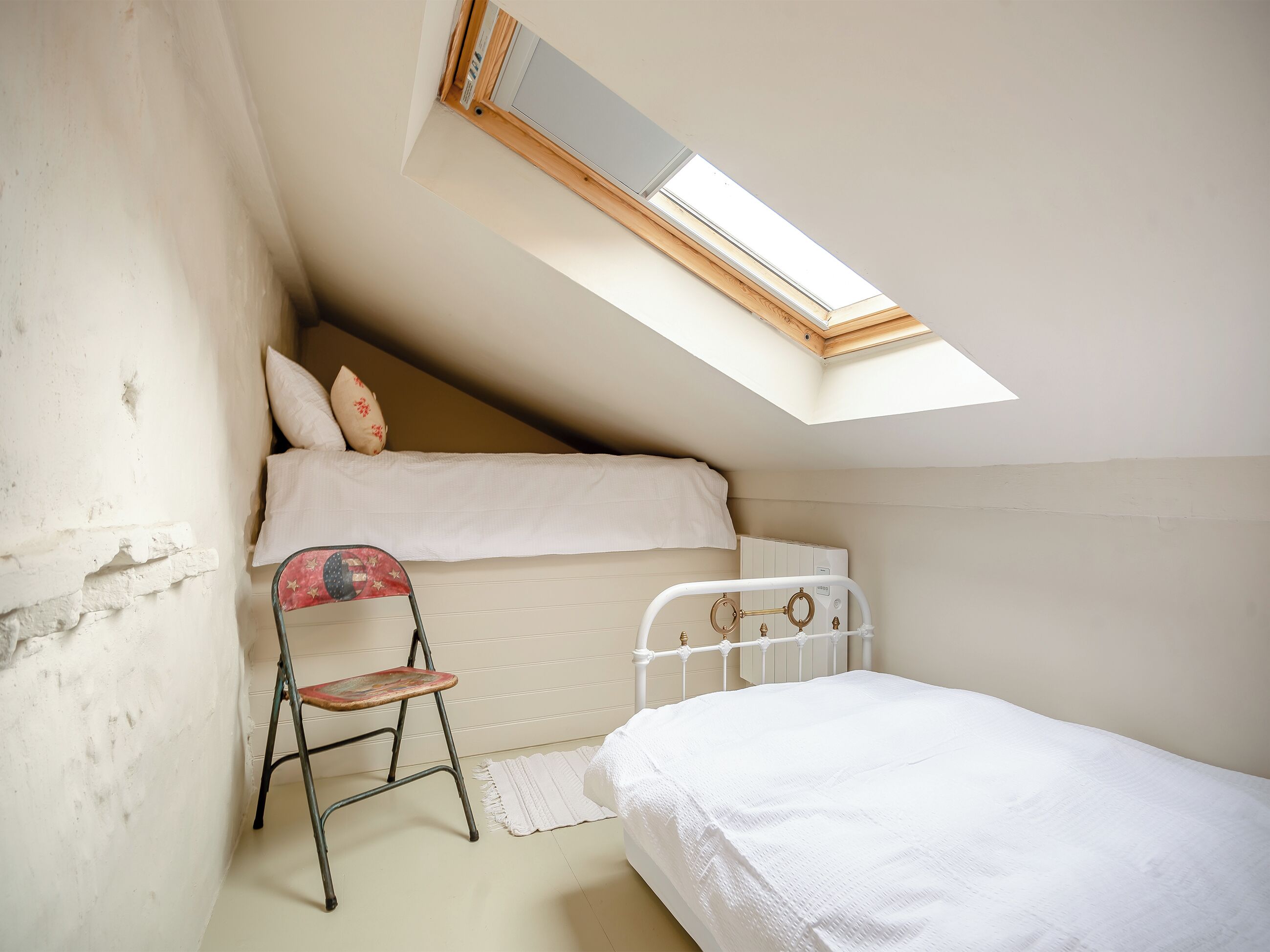 A dormitory room with a bunk bed, a small chair, a skylight, and a radiator.