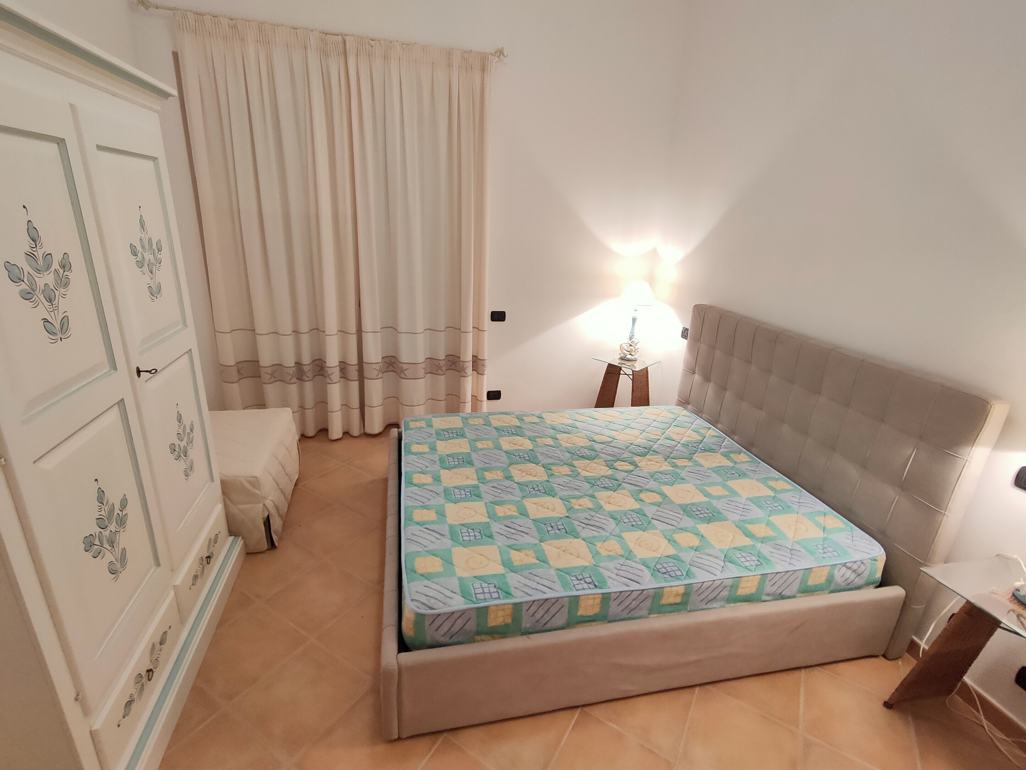 A bedroom with a bed, a nightstand, a lamp, a wardrobe, and a window with curtains.