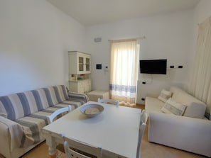 Una sala de estar con un sofá a rayas, un sillón blanco, una mesita con un cuenco, un aparador y un televisor montado en la pared.